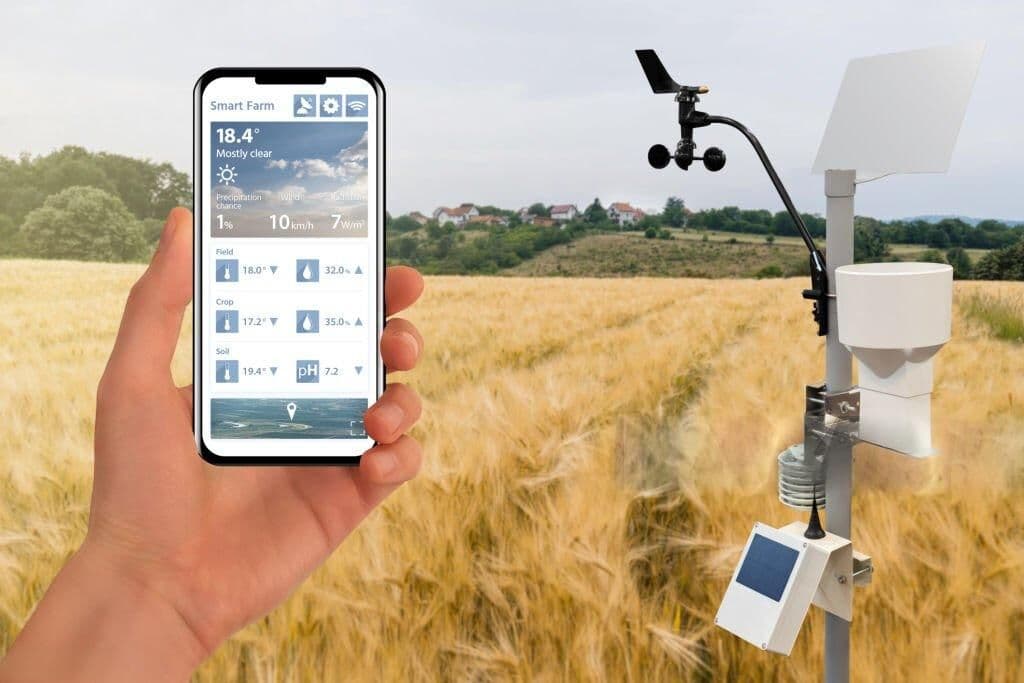 Hand holding a phone showing a Smart Farm weather and soil advisory app, with a field weather station in the background