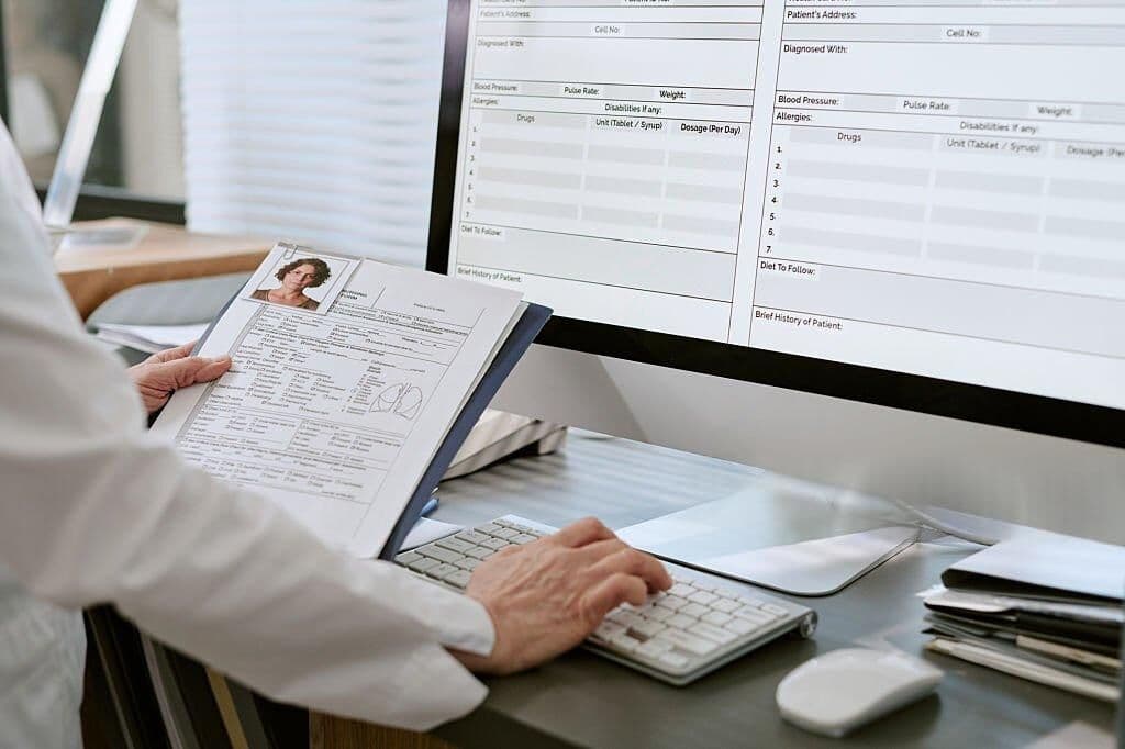 Clinician reviewing a patient chart on screen alongside a printed patient file, generic EHR layout with no real patient data