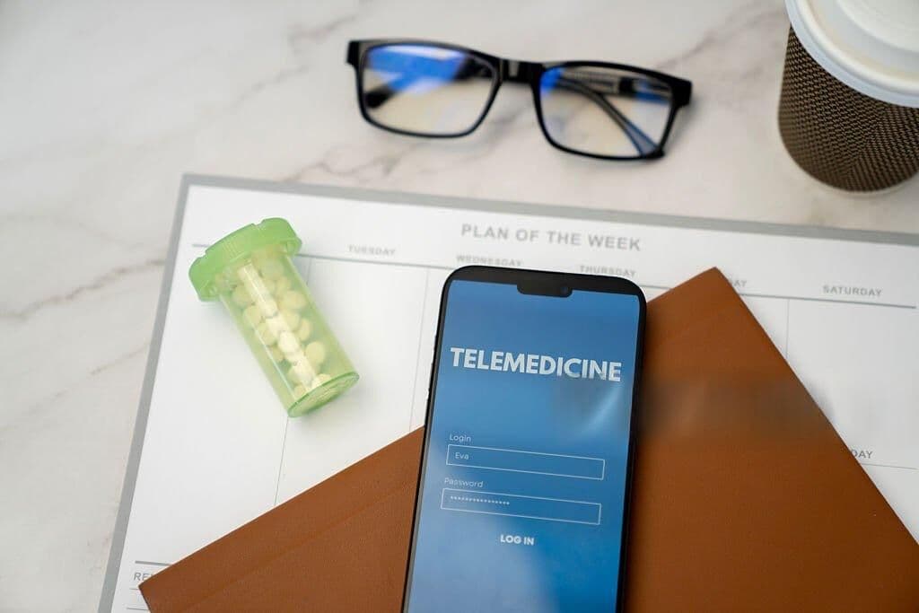 Phone showing a telemedicine app login screen on a desk with glasses and a prescription bottle